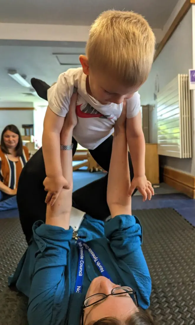 A little boy balances on his key worker’s raised feet, flying like an aeroplane.