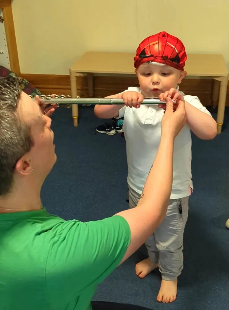 A little boy in a red hat reaches up to blow into a flute that Kate is holding for him.