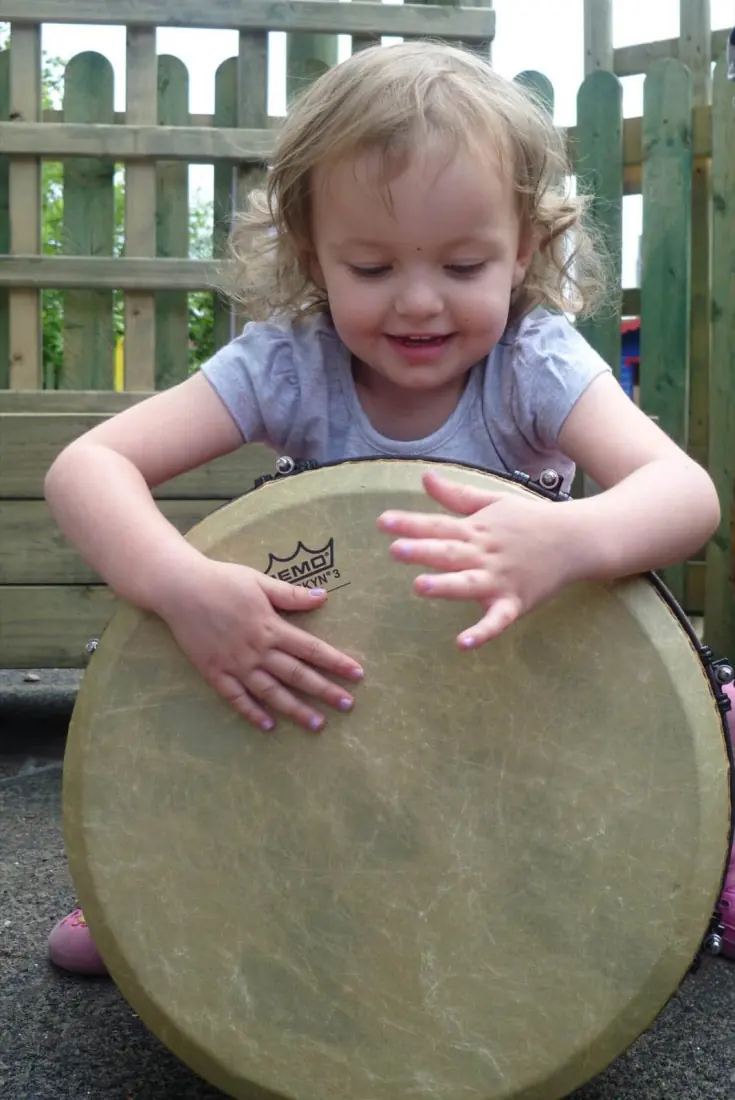 A smiling toddler leans over a big djembe drum, both hands on the skin.