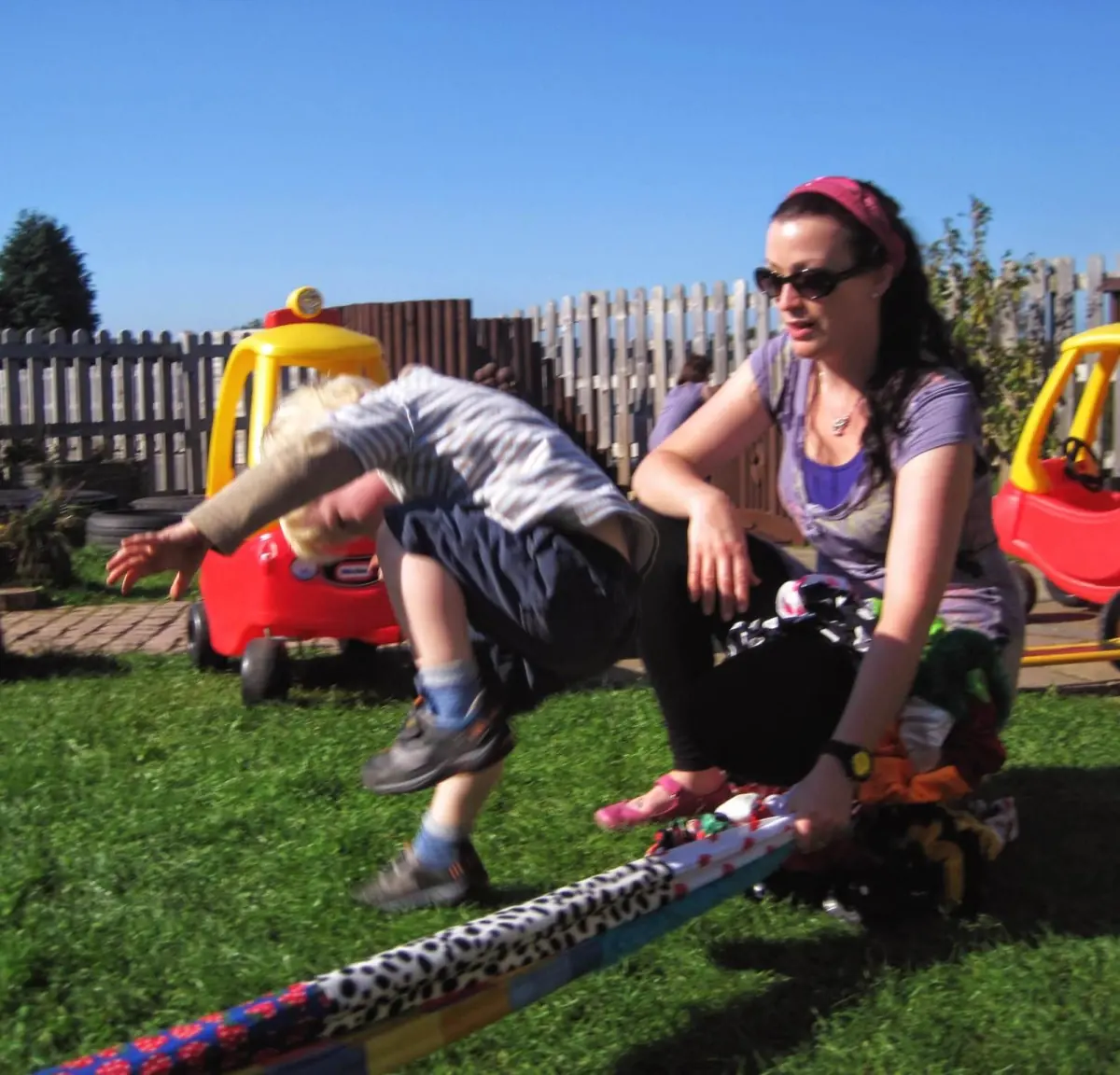 A child leaps over a colourful scrunchy on the grass while a grown-up holds it low.