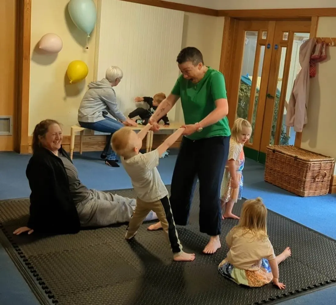 Kate holds a boy’s hands as they move together, with children and grown-ups playing around them.