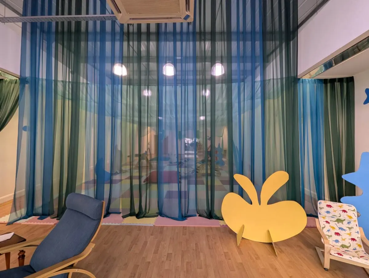 Inside Cocoon: blue and green voile curtains hang from the ceiling over pastel floor tiles, with a cut-out yellow splash shape standing on the wooden floor.