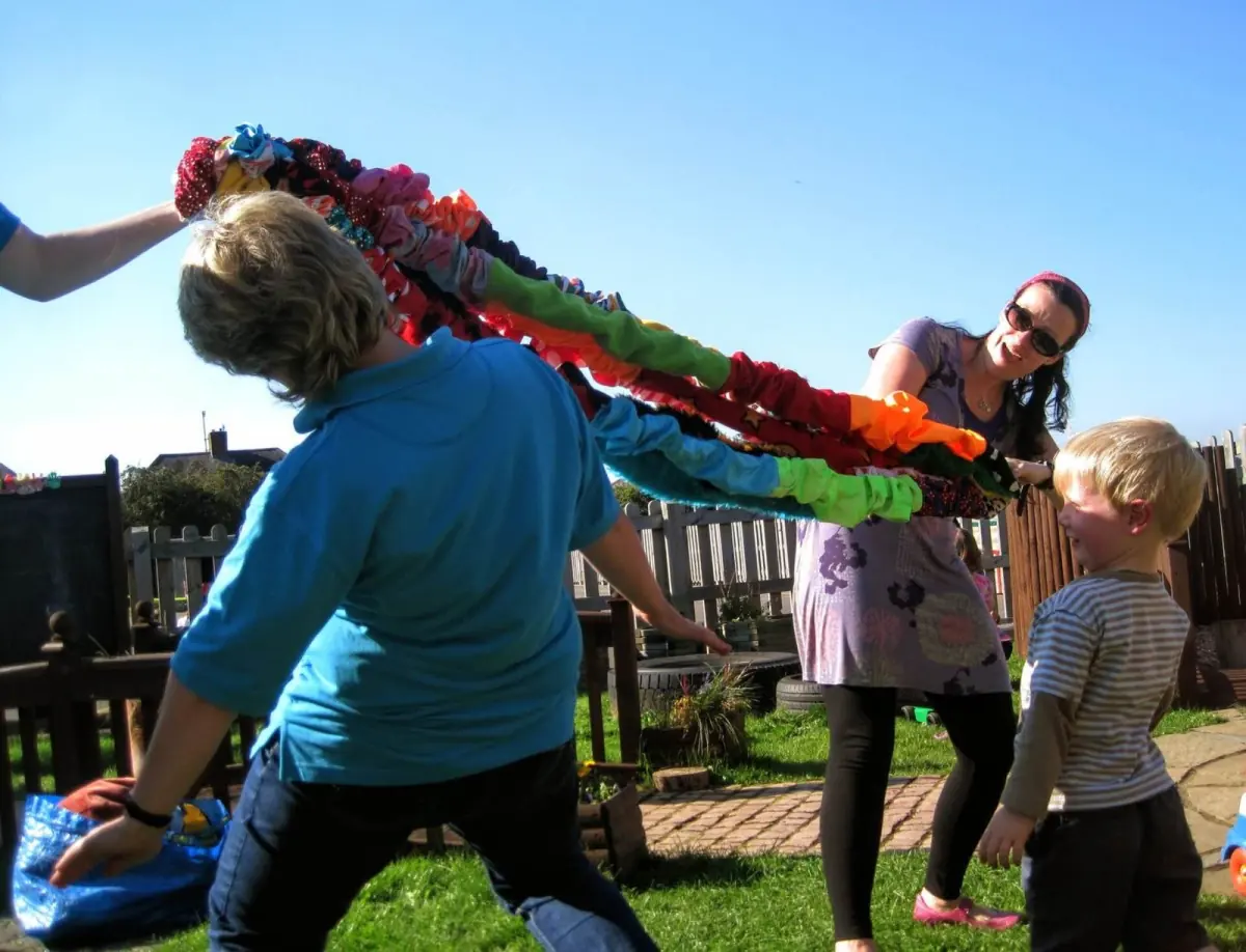 Grown-ups and children on the grass in the sunshine, lifting a long, colourful scrunchy high.