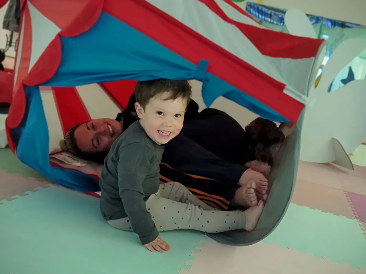 A grinning toddler crawls out of the red and blue play tent at Cocoon, with his mum Megan, Cocoon's co-director, lying in the tent behind him.