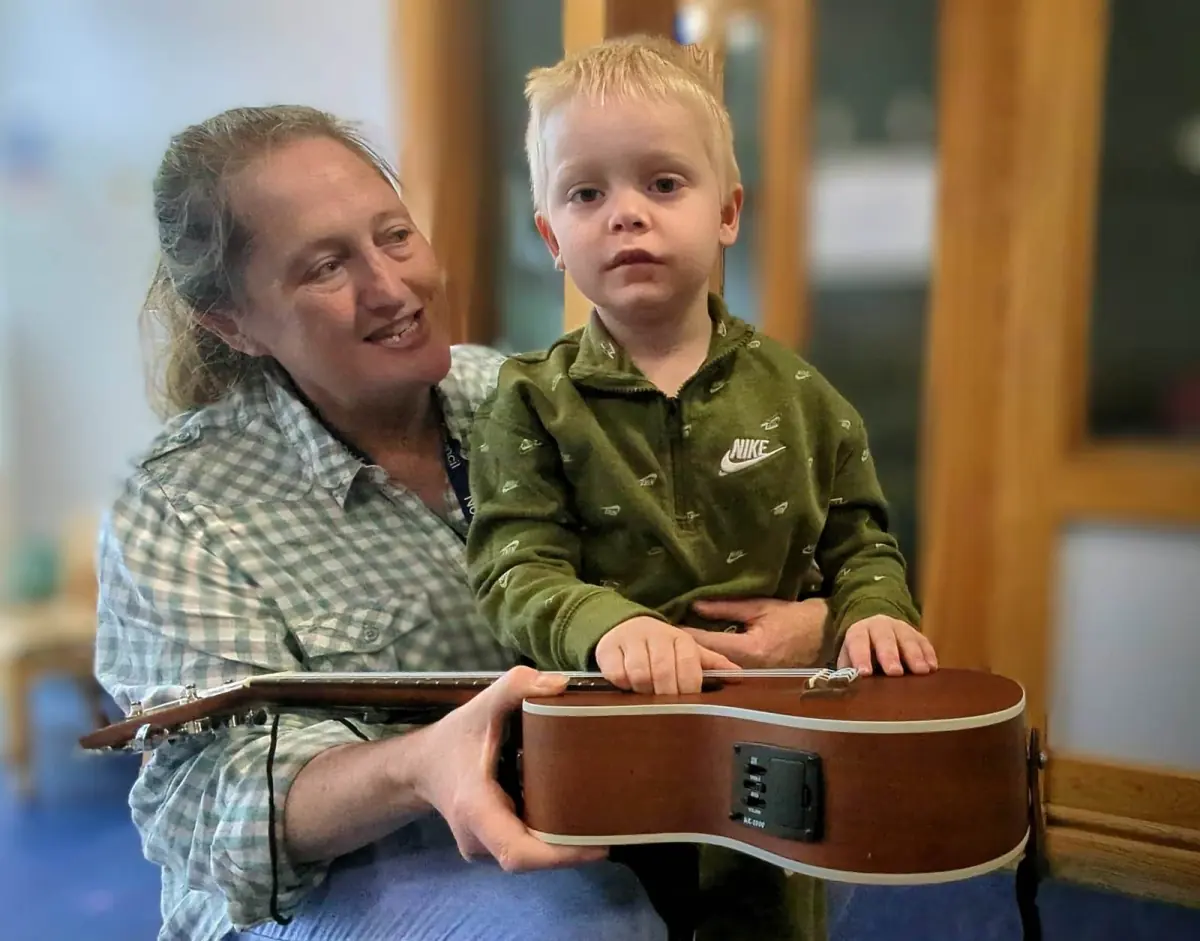 A boy holds the ukulele strings, sat on his key worker’s knee.
