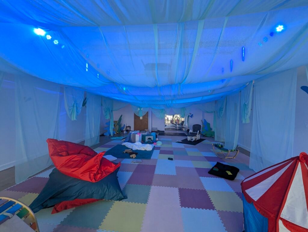 The Cocoon playspace, lit soft blue, with a duvet made up like a bed on the floor, a big bean bag, a play tent and soft foam flooring.